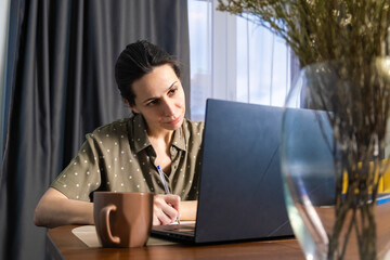 Focused woman taking handwritten notes with a pen while engaging with content on a laptop screen, illustrating effective hybrid learning, online education, or detailed study from a home office
