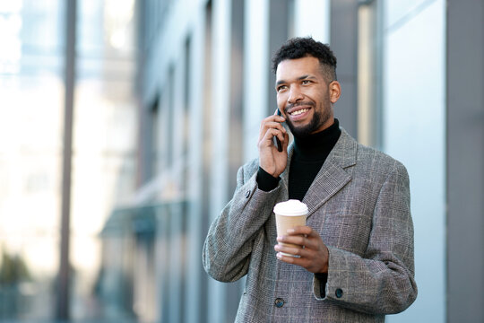 Smiling man in suit with takeaway cup talking by smartphone on city street, low angle view. Space for text