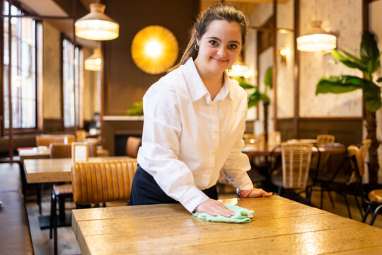 A young girl with Down syndrome works as a waitress in a modern cafe wearing a uniform.The girl looks at the camera while cleaning a table.Concept of trisomy 21. - Powered by Adobe