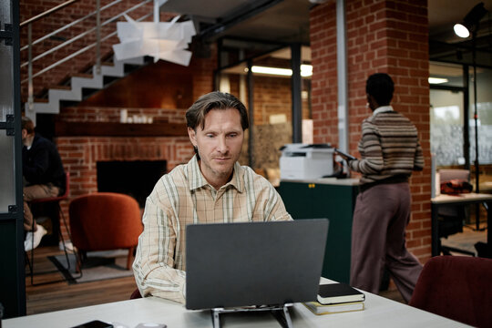 Caucasian middle aged man working on laptop at desk in modern business office while Black woman standing near printer in background, both focused on professional tasks, open workspace visible