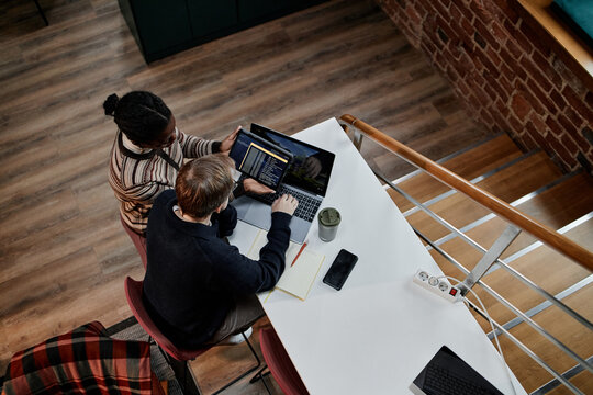 Young adult Black woman and young adult Caucasian man collaborating on laptop at modern business office desk, both focusing on computer screen with smartphone and notebook nearby
