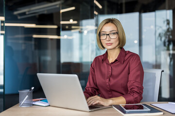 Professional businesswoman wearing glasses sitting at an office desk, typing on a laptop, and looking confidently at the camera, embodying dedication and corporate success in her modern workspace