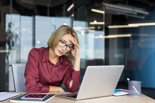 Businesswoman experiencing headache and stress at her modern office workspace, feeling overwhelmed and tired while working on her laptop, depicting concepts of burnout and work pressure