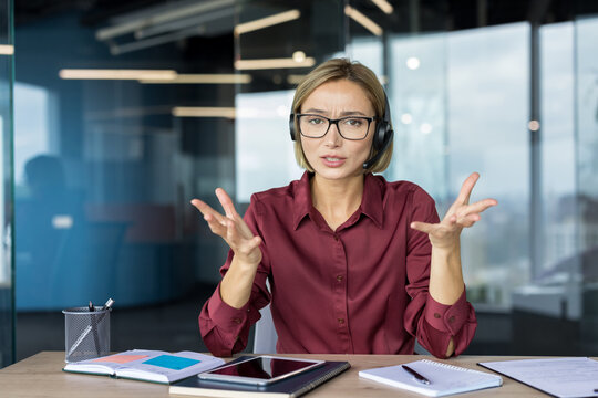 Businesswoman wearing a headset engaging in a video call, expressing confusion and frustration with open hands while working at her office desk in a modern workplace