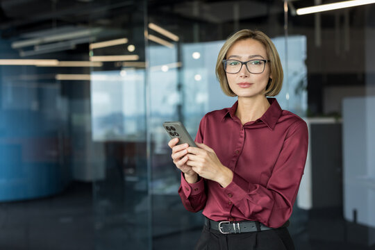 Professional asian businesswoman standing in modern office holding a smartphone, looking at camera while engaging in communication, work, or social networking