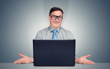 Confused businessman in glasses and tie sits at laptop, shrugging with hands spread, showing frustration or helplessness over a problem on computer.