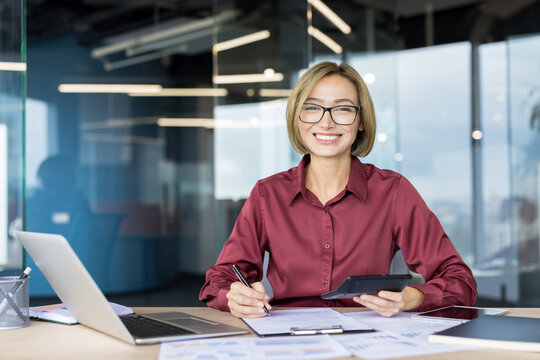 Young female professional accountant working with documents and a calculator at her desk, smiling confidently at the camera, representing success, financial planning, and business management - Powered by Adobe