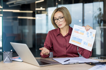 Businesswoman discussing business strategy and financial data, holding a document with graphs during a remote video conference meeting using her laptop in a modern office