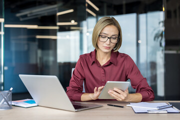 Confident businesswoman multitasking at a desk, using laptop and tablet to manage digital communication and strategy in a modern corporate office environment