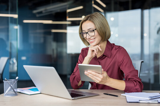 Young businesswoman with glasses smiling, interacting with a digital tablet and laptop at her modern office desk, demonstrating productivity and technology integration