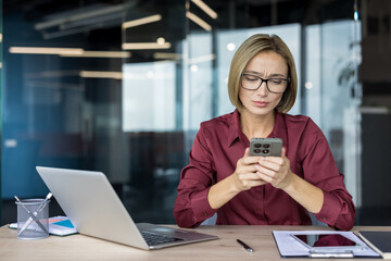 Businesswoman sitting at her office desk, holding a mobile phone and looking at the screen with a stressed, concerned expression, reacting to problematic information or bad news