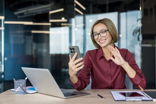 Businesswoman cheerfully participating in a video call on her smartphone in a modern office, waving her hand while communicating with colleagues or clients