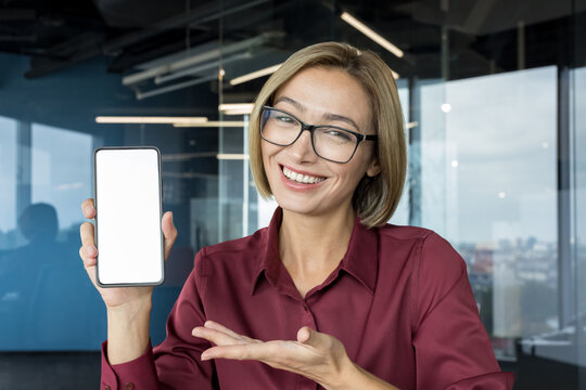 Female professional entrepreneur enthusiastically showing a mobile phone with a white empty screen for advertising or app promotion against a modern office background