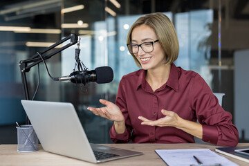 Asian woman recording or streaming online podcast, speaking into a professional microphone and...