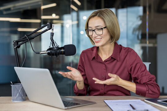 Asian woman recording or streaming online podcast, speaking into a professional microphone and looking at a laptop while gesturing in a modern office, showing broadcasting and communication concepts - Powered by Adobe