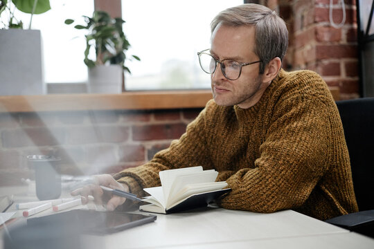 Middle aged Caucasian man wearing glasses working at desk in modern business office, holding pen and writing in notebook while looking at digital tablet, brick wall visible in background