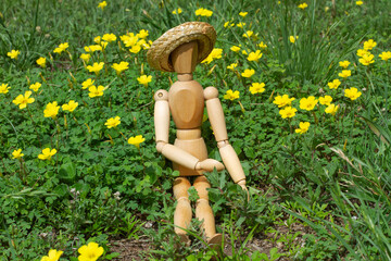 A wooden mannequin toy wearing a wicker hat sitting on the grass with yellow flowers around