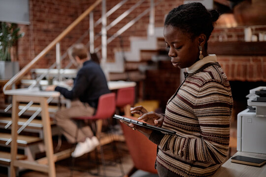 Young adult Black woman using digital tablet near printer in modern business office while middle aged Caucasian man working at desk in background, both focused on professional tasks