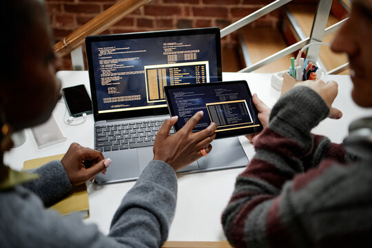 Young adult Black woman and young adult Caucasian man collaborating on software development project, reviewing code displayed on laptop and digital tablet in modern business office