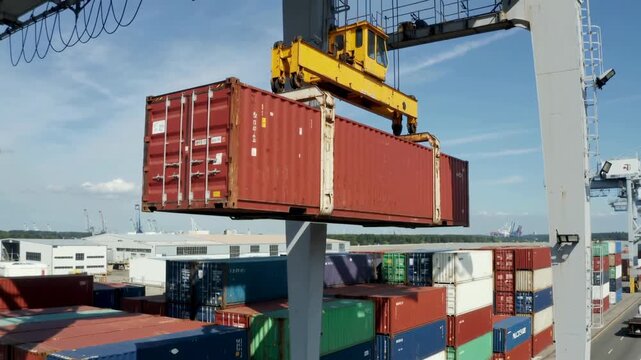 The video shows a large red cargo container being lifted by a yellow crane in a port or shipyard. Other containers are visible in the background, indicating a busy cargo loading and unloading area. Th