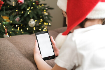 Five-year-old boy in Santa hat using a smartphone with blank white screen on sofa next to decorated Christmas tree. Christmas holidays, video calls, mobile apps, screen mockup