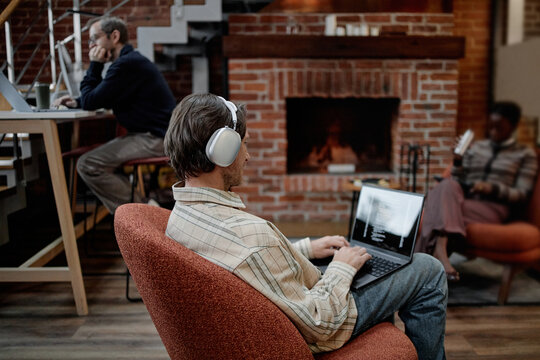 Caucasian young adult man wearing headphones working on laptop in modern business office, middle aged Caucasian man using desktop computer in background, Black woman sitting on chair