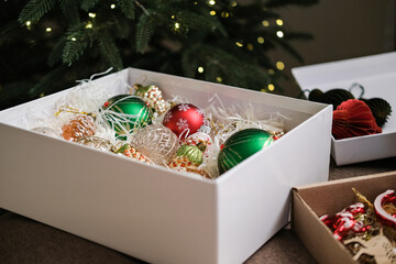 Close-up of white box filled with red, green and clear glass Christmas ornaments, next to fir branches and other decorative boxes. Christmas ornament storage, decorating, holidays, gift wrapping
