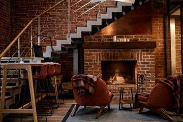 Fototapeta premium Modern business office interior featuring empty seating area with mid century style chairs and barstools arranged near brick fireplace and staircase, no people visible