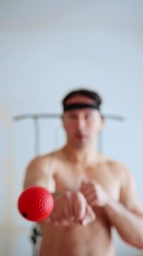 bare-chested man mastering reflex boxing drill, casual home session with red elastic ball, orange shorts, headband, quick hand movements and playful intensity, focus on reaction timing and technique.