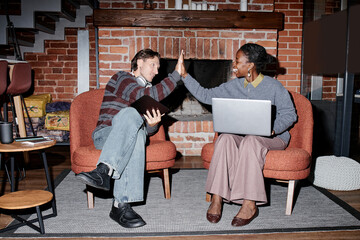 Young adult Caucasian man and middle aged Black woman sitting in modern business office high fiving while holding laptop and tablet, both smiling and engaging in collaborative teamwork