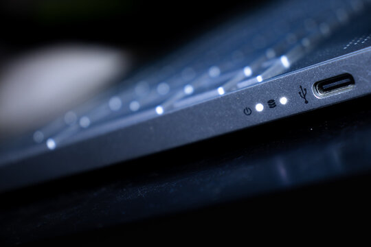 The slim profile of a laptop is displayed in the dark, with white LED status indicators for power and storage disk activity clearly illuminated near a USB-C port.