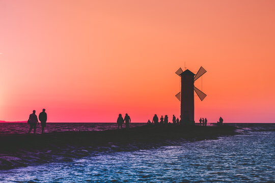 A group of people walk along the pier near the lighthouse on the sea at sunset