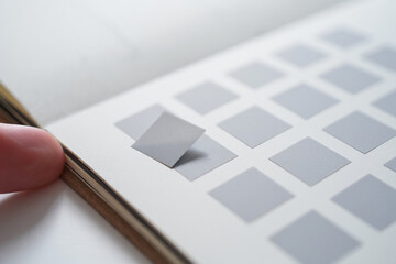 Hand Placing Small Gray Square Sticker Into a Book of Blank Squares for Arts and Crafts on Bright Surface with Light