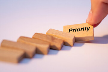 Hand Placing Wooden Block Marked Priority on Top of Others Demonstrating Importance Concept Close up Shot with Selective Focus on White Background
