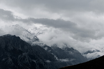 Snow-capped mountains rise above a dark, forested landscape under a cloudy sky. The scene captures the beauty of nature in a dramatic setting.