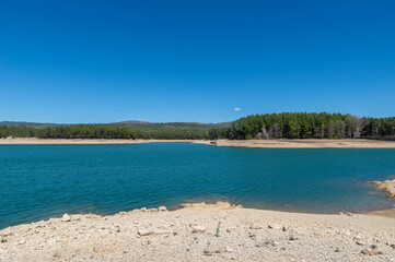 Bright blue waters gently lap against the shore where pebbles and sand meet the edge of a tranquil lake surrounded by lush green trees. The scene evokes peace and natural beauty