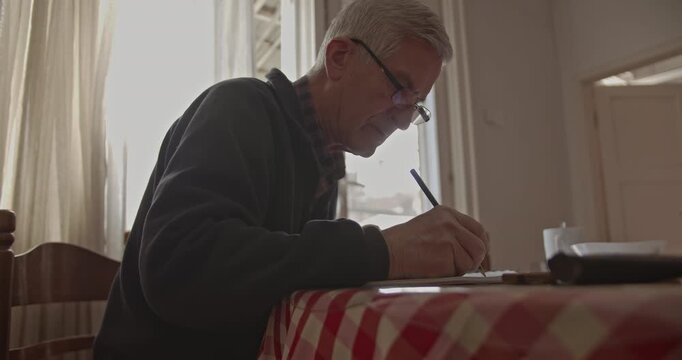 An older man, focused and engaged, carefully writes on paper before switching to his smart phone at a table in his quiet home. This captures a moment of domestic activity.