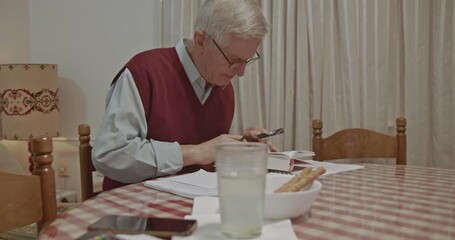 An elderly senior man is intently reading a book with a magnifying glass at home, diligently taking notes on paper. He is focused on studying, learning, and managing important documents and paperwork.