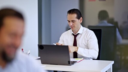 Focused professional businessman works on his laptop and engages in a phone call simultaneously at his office desk. He smiles, showcasing efficiency and successful multitasking in a corporate setting. - Powered by Adobe