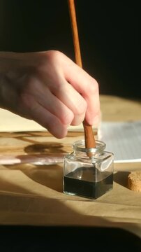 Close up writer hand dipping classic wooden dip pen with metal nib into glass inkwell of black ink preparing to write on aged paper in warm natural sunlight. Calligrapher dipping vintage pen. Vertical