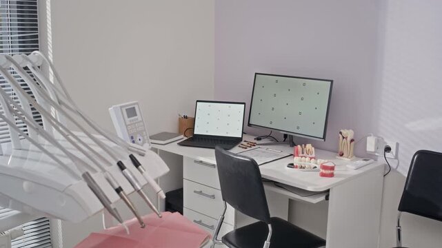 No people shot of bright modern dental office featuring treatment chair unit and dentist desk with desktop, laptop, and prosthetic teeth samples, ready for patient care
