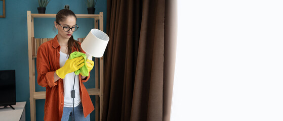 Woman wearing yellow gloves cleaning a white table lamp with microfiber cloth in the living room near window. Banner. Household, housekeeping and house cleaning