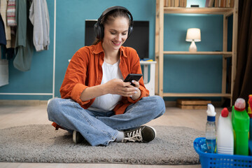 female janitor with Cleaning supplies using smartphone and headphones sitting resting on floor at home