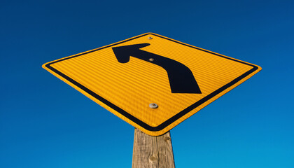 Yellow diamond road sign with a black curved arrow pointing left against a clear blue sky.