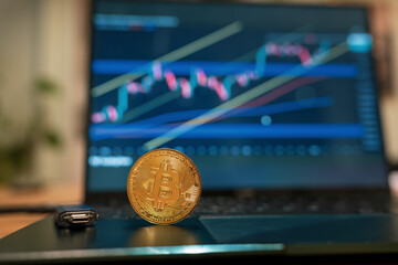Close up of a physical bitcoin coin standing on a laptop keyboard with a wallet and financial trading charts displayed on the screen behind