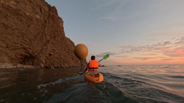 Kayaking ocean sunset person paddling kayak with large balloon near coastal cliffs beautiful outdoor activity