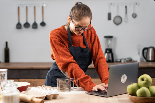 Happy pastry chef woman typing using laptop at cozy kitchen