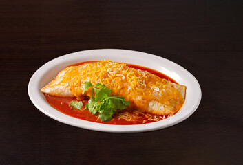 Jumbo burrito covered with melted cheese garnished with parsley in tomato sauce in a white bowl on a dark surface; copy space