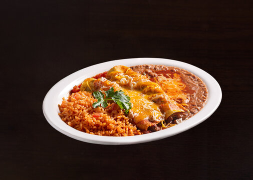 Enchilada platter with rice and refried beans covered in melted mozzarella cheese and a sprig of parsley in a white plate; copy space