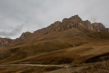 Panoramic view of the mountainous landscape in Kabardino-Balkaria. Rolling hills with dry grass and distant snow-capped peaks under a cloudy sky.